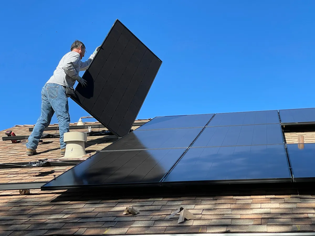 A solar installer lifting a black panel into place on a tiled roof under a clear blue sky